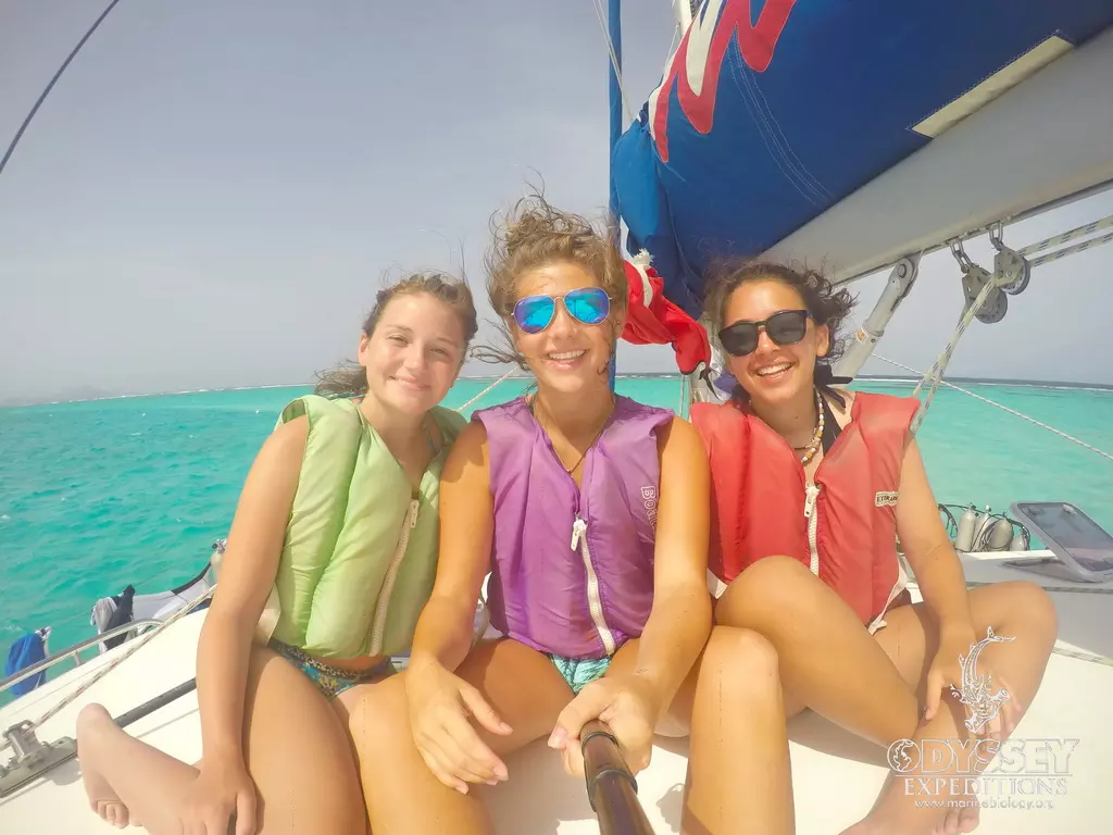 Three Odyssey teens on the catamaran bow with the boom sail behind them, sailing through clear Caribbean water